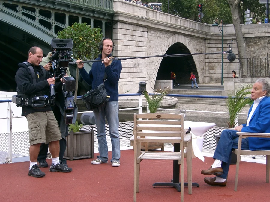 Équipe de tournage en extérieur, caméra et perche au-dessus d'une personne interviewée au bord de la Seine